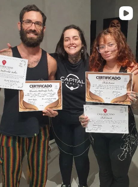 Nina Piercer com dois alunos formados, segurando os certificados do curso de Body Piercing no Estúdio Aurya
